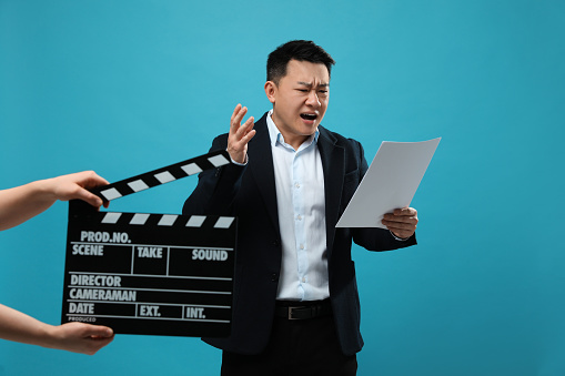 Emotional asian actor with script performing while second assistant camera holding clapperboard on light blue background. Film industry