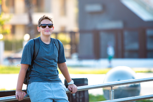 Radiating youthful energy, a stylish teen boy wearing sunglasses stands confidently in front of the school, eagerly awaiting the start of classes on a bright and sunny day. Dressed in smart gray casual attire, he exudes a happy and cool demeanor, blending a sense of style with a readiness for learning. This image captures the essence of a carefree and smart approach to school life, where the sunlit ambiance complements the teen's enthusiasm for the day ahead. A perfect blend of casual charm and academic coolness in a vibrant school setting.