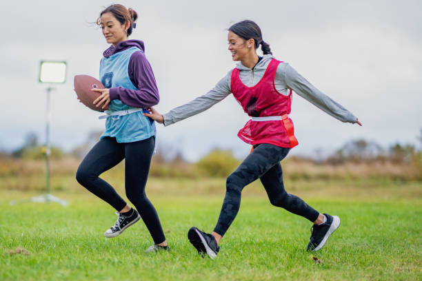 Two University students are seen running on a field as they compete on opposite teams during a flag football game.  They are both dressed comfortably and are wearing numbered team pinnies.