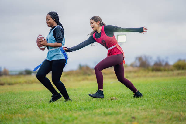 Two University students are seen running on a field as they compete on opposite teams during a flag football game.  They are both dressed comfortably and are wearing numbered team pinnies.