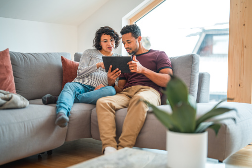 Cute mid adult multicultural pregnant couple sitting in a living room, using a digital tablet. They are surfing the net.