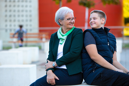 In a modern and vibrant public space, a teen in a stylish dark blue jacket and his elegant grandmother, adorned in a dark green coat, bask in the warmth of a sunny day. Without the distraction of screens, they immerse themselves in the simple joy of each other's company, creating lasting memories amidst the contemporary backdrop of a red building. This heartening image captures the essence of intergenerational connection, showcasing the beauty of shared moments and genuine happiness in the embrace of family love.