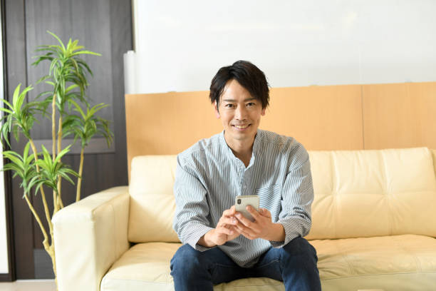 istock Smiling young Asian man sitting on the sofa while holding a mobile phone 1792098264