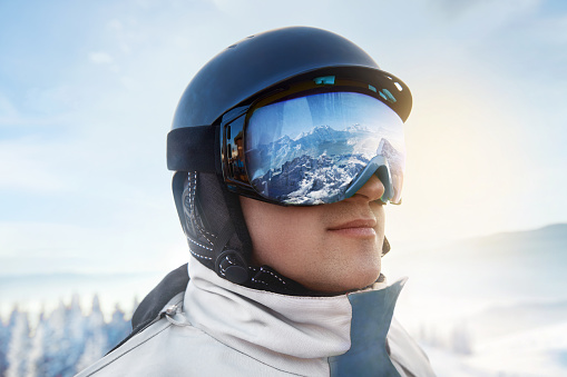 Close Up Of The Ski Goggles Of A Man With The Reflection Of Snowed Mountains. Man In The Background Blue Sky.  Winter Sports