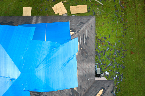 Aerial view of damaged in hurricane Ian house roof covered with blue protective tarp against rain water leaking until replacement of asphalt shingles.