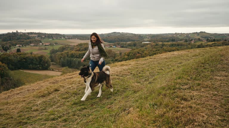 SLO MO Cheerful Woman and Cute Akita Dog Running on Grassy Field Under Cloudy Sky in Countryside