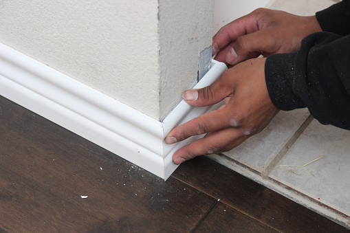 Hands carefully installing white baseboard at the corner of a wall