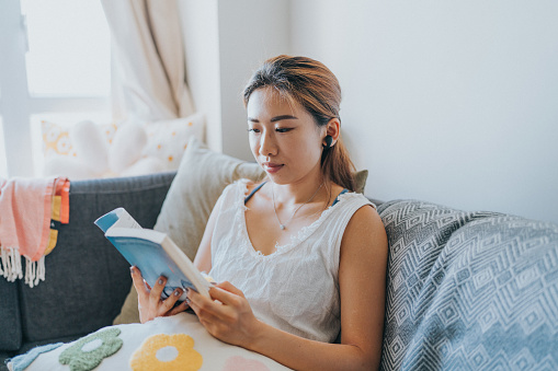 A medium shot of a young East Asian woman with brown hair reading a blue novel on her sofa. She is wearing a white top and earphones. She feels relaxed and comfortable with a small cushion on her lap and back. The living room is lively as the sun shines brightly passing through the window.