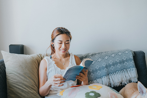 A medium shot of a young East Asian woman with brown hair reading a blue novel on her sofa. She is wearing a white top and earphones. She feels relaxed and comfortable with a small cushion on her lap and back. The living room is lively as the sun shines brightly passing through the window.