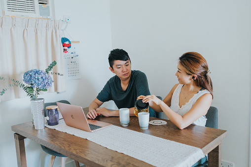 A wide shot of a young East Asian couple working on a table using a laptop while enjoying tea in the morning.