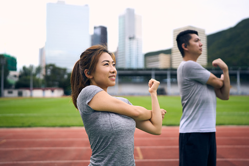 A medium shot of a Chinese couple doing arm stretches on the sports ground on a sunny day. They are exercising near a running track with buildings and grasses in the background. The photo only showcases half of their bodies.