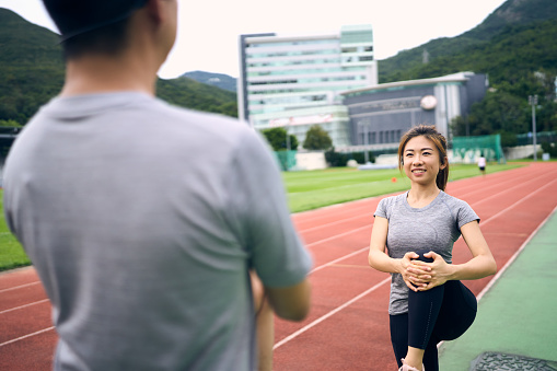 A medium shot of a Chinese couple doing leg stretches on the sports ground on a sunny day. They are exercising near a running track with buildings and grasses in the background. This photo focuses on the woman.