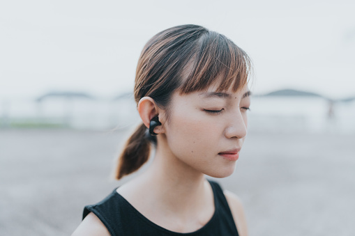 A close-up of a young East Asian female closing her eyes and listening to music. She has brown hair and wears a black tank top. She is relaxing herself before she goes jogging. She is in a park near the city with a lively atmosphere and sunny weather.