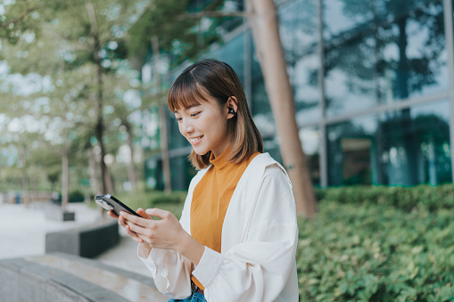 A medium shot of a young East Asian female adult looking and using her mobile phone. She is sitting in an open space surrounded by trees and bushes near her office. She is wearing an orange shirt, jeans, and white jacket.