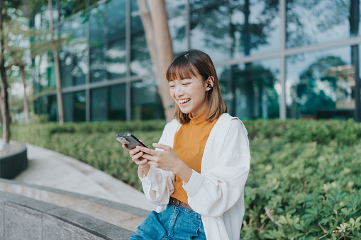 A medium shot of a young East Asian female adult looking and using her mobile phone while laughing. She is sitting in an open space surrounded by trees and bushes near her office. She is wearing an orange shir, jeans, and white jacket.