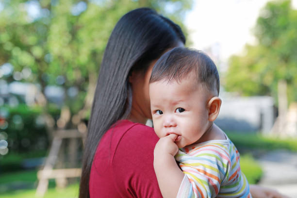 portrait d’un bébé garçon suçant le doigt avec une mère asiatique portant. - enfold photos et images de collection