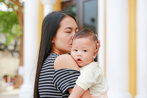 Portrait of Asian mother carrying and kissing her infant baby boy outdoor.