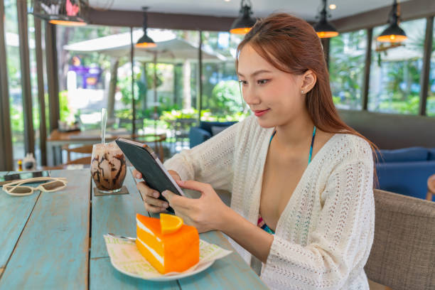 istock Young asian woman using a digital tablet while having an iced latte and orange cake in a beach cafe. Female digital nomad wearing beachwear using tablet in vacation vibes. Asian woman in beachwear remote working in a cafe. 1792322073