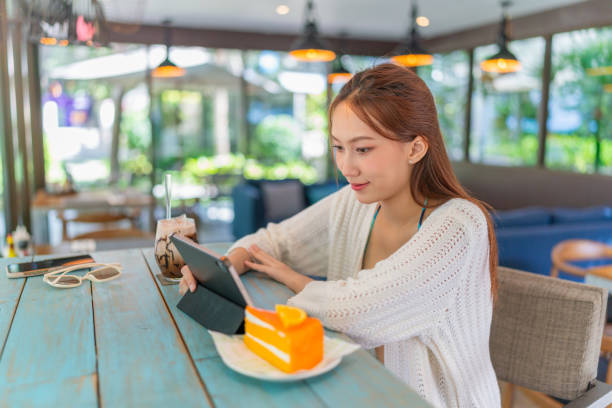 istock Young asian woman using a digital tablet while having an iced latte and orange cake in a beach cafe. Female digital nomad wearing beachwear using tablet in vacation vibes. Asian woman in beachwear remote working in a cafe. 1792322584