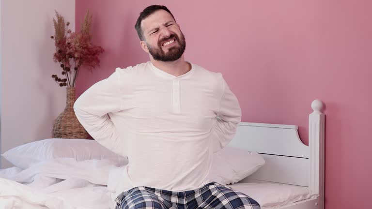 Young bearded man suffering for backache sitting on bed at bedroom