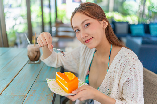 Young asian woman enjoying a slice of orange cake while having an iced latte in a beach cafe. Female digital nomad wearing beachwear taking a break in vacation vibes. Asian woman in beachwear relaxing in a cafe.