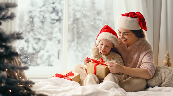 Merry Christmas and Happy Holidays. Cheerful mom and her cute daughter girl exchanging gifts. Parent and little child having fun near tree indoors. Morning Xmas.