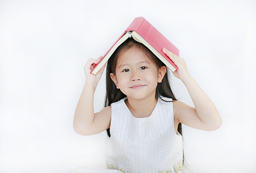 Portrait of little Asian girl place hardcover book on her head and looking camera over white background.