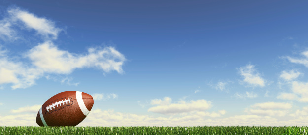 American football ball, on the grass, with fluffy couds sky at the background. Side view, from ground level, panoramic format.