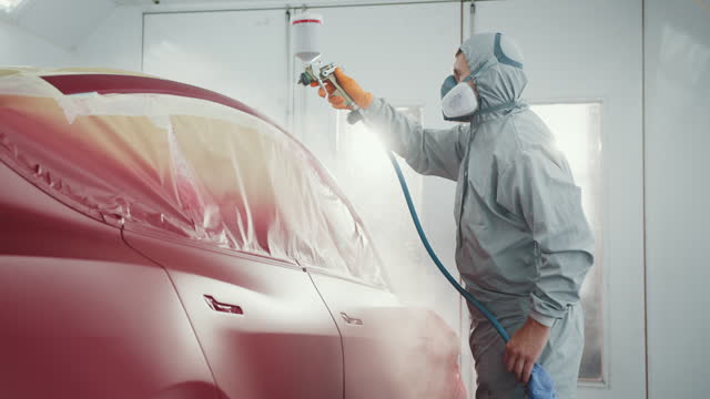 Male Painting Technician Applying Red Paint to a Car Part Spray Gun in a Car Body Shop. Car Service Station. Worker Painting a Red Electric Car in Special Garage, Wearing Costume and Protective Gear.