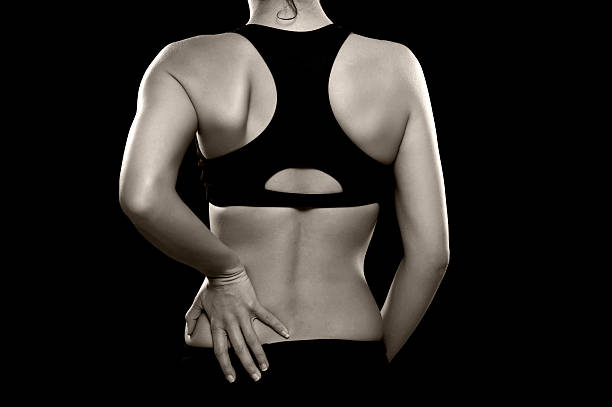 Woman with Lower Back Pain A black and white photo of an athletic woman holding her lower back as if experiencing pain. Photographed in the studio. Isolated on a black background. coccyx photos stock pictures, royalty-free photos & images