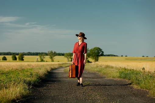 Girl in red dress with suitcase on country road in sunset