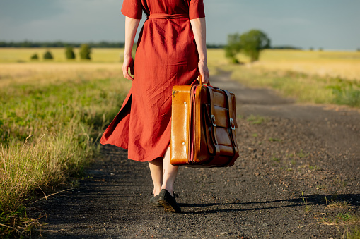 Girl in red dress with suitcase on country road in sunset. Low side view