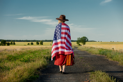 Girl in USA flag with suitcase on country road in sunset