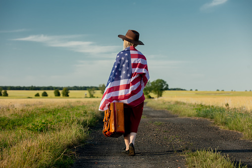 Girl in USA flag with suitcase on country road in sunset