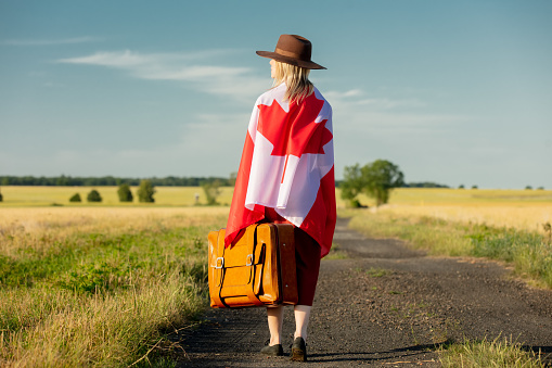 Girl in Canada flag with suitcase on country road in sunset