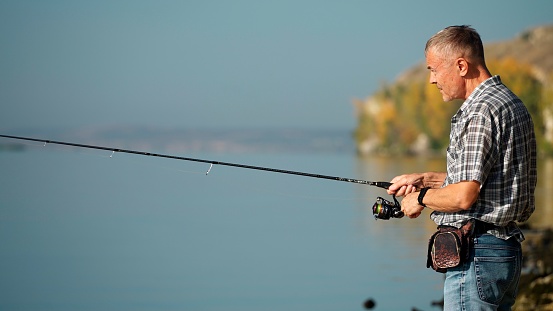 In autumn, a mature adult man in a shirt and jeans catches fish with a spinning rod on the river bank. Senior adult goes fishing on weekends. A young pensioner is engaged in a hobby.