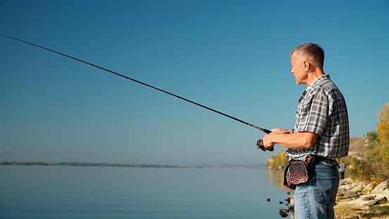 Profile view of an adult male fisherman in a shirt and jeans fishing on the river bank with a spinning rod. Mature european man enjoys his favorite hobby on a sunny autumn day on the weekend