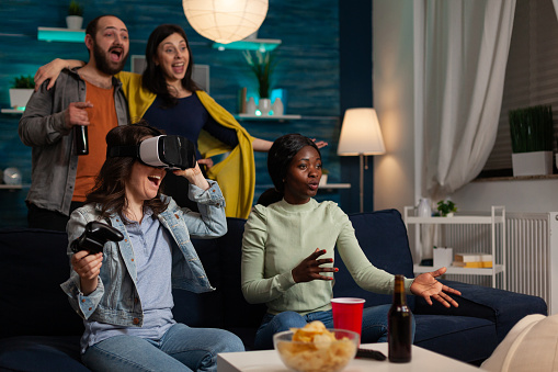 Excited happy woman with virtual reality headset winning gaming competition playing online videogames using joystick enjoying victory with friends at home. Concept of multu-ethnic friends hanging out