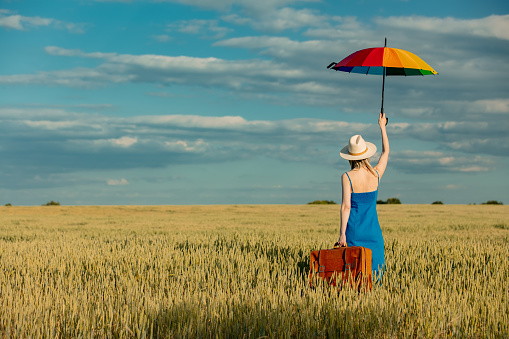 Girl in blue dress with suitcase and umbrella in wheat field