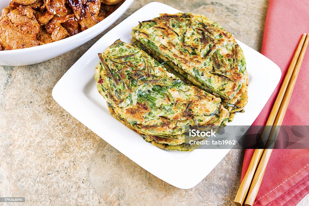 Table set for Korean Green Pancakes and barbequed pork Horizontal photo of Korean green onion pancakes in plate, chopsticks on cloth napkin, and spicy barbequed pork in bowl on stone table Korea Stock Photo