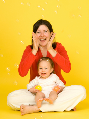 Happy and fun mother daughter portrait for Spring. Baby girl sitting on mother's lap and grinning from ear to ear while mom claps, adding to the excitement of many bubbles floating in the air. Yellow background.