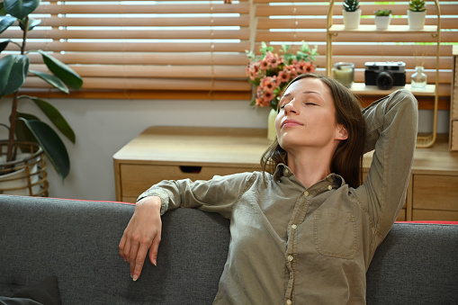 Calm young woman with hand behind head, closing eyes relaxing on comfortable sofa at home.