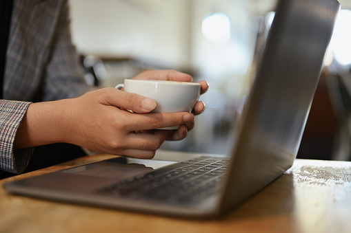 Unrecognizable businesswoman holding cup of coffee using laptop on office desk.