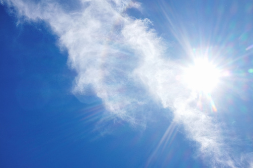 Majestic beauty of the sky with a focus on a white, fluffy cloud that is highlighted by radiant sunbeams piercing through it. The background is a vibrant blue. Lens flare creates rays coming from the direction of the sun. A serene depiction of natural daylight beauty, evoking a sense of tranquility and the sublime in nature.