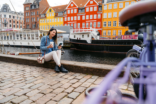 Beautiful young woman using her smart phone while drinking coffee in Nyhavn Canal in Copenhagen in Denmark.