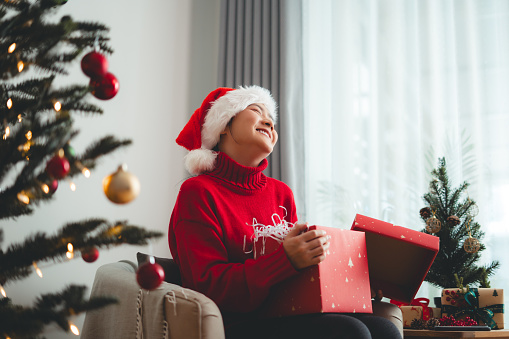Asian woman having fun opening gift boxes on Christmas day, sitting in living room decorated with a Christmas tree.