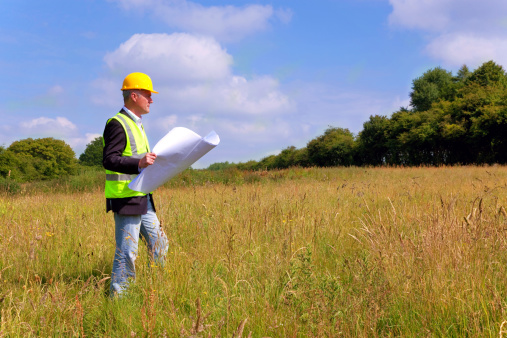 Architect wearing site safety gear and holding a set of plans whilst surveying a new building plot