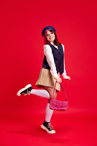 Young girl, student with bright makeup and piecing, in stylish clothes, cheerfully posing against red studio background. Concept of youth culture, self-expression, education, fashion, emotions