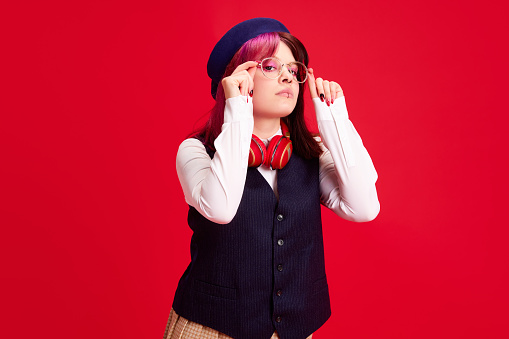 Young girl, student with pink hair and piercing, wearing stylish clothes with beret, standing against red studio background. Concept of youth culture, self-expression, education, fashion, emotions