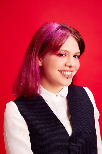 Portrait of young girl with pink hair, bright makeup and piecing smiling, looking at camera against red studio background. Concept of youth culture, self-expression, education, fashion, emotions